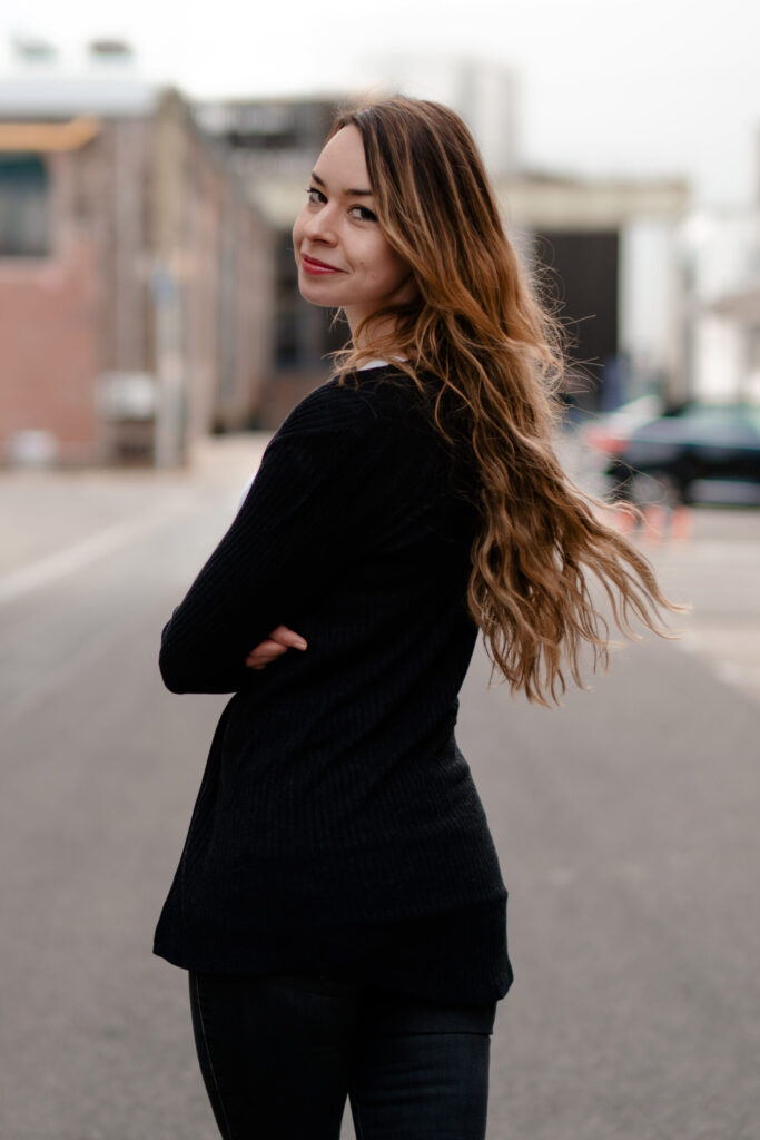 Person mit langen Haaren blickt über die Schulter in die Kamera, aufgenommen im Freien. - Datingfotos Köln