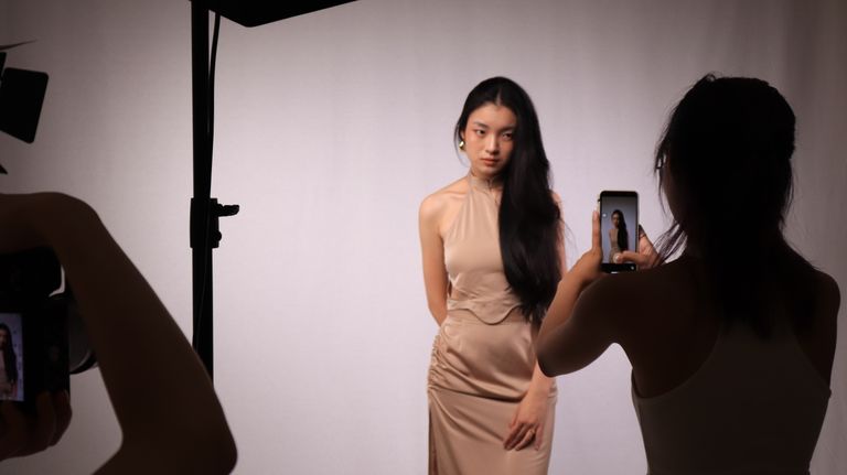 A professional studio photoshoot capturing a woman in a beige dress, using digital cameras and smartphones.